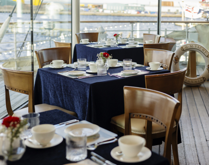 There are tables set with blue tablecloths and tea settings in the Royal Deck Tearoom. Through the windows you can see the Port of Leith. 
