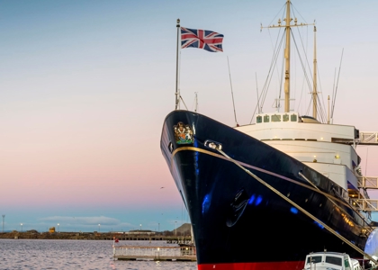 A large, elegant ship is docked at the harbour at sunset, its dark blue hull and white superstructure reflected in the calm water. A Union Jack flies at the bow, with smaller boats moored nearby and warm evening light across the scene.