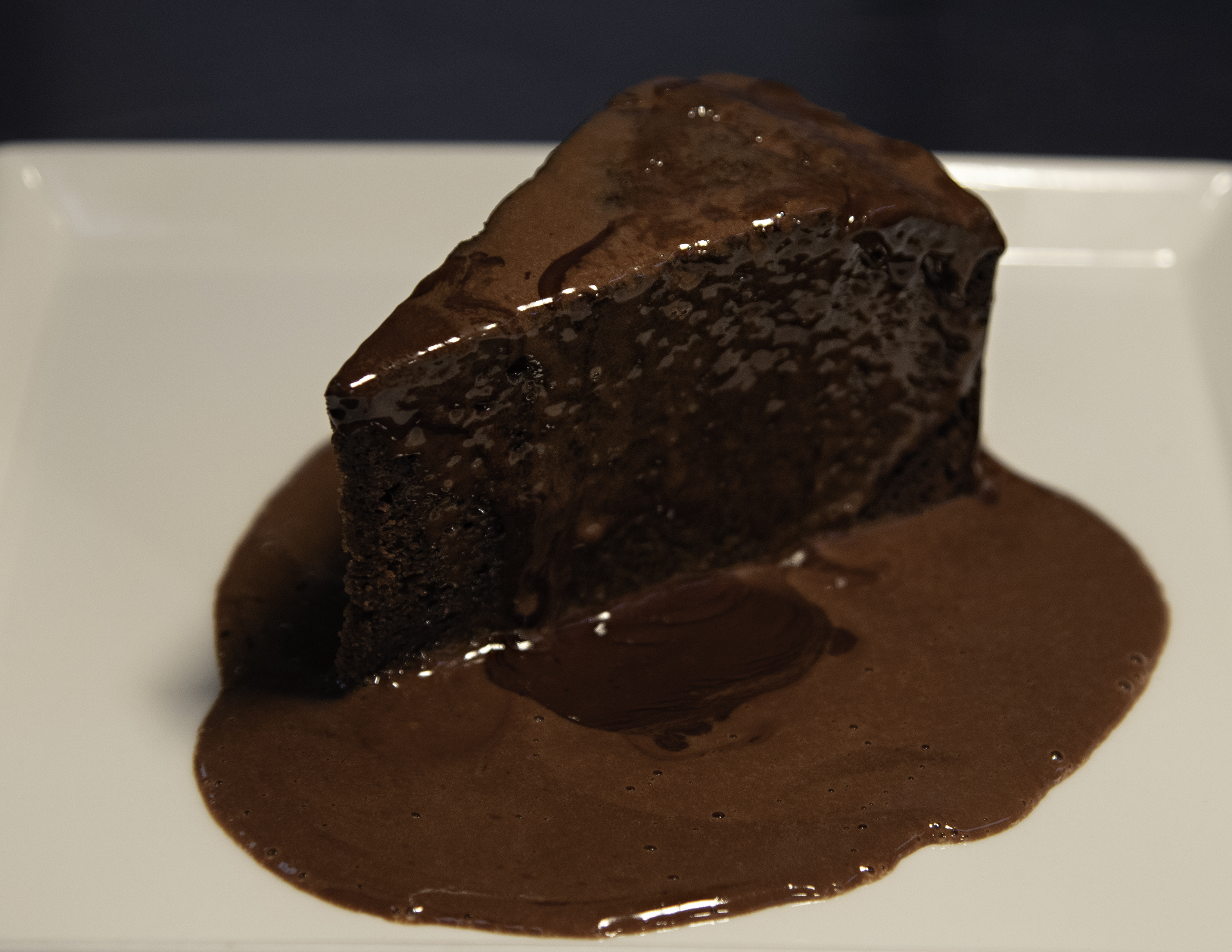 A slice of chocolate fudge cake with melted chocolate sauce on a plate.