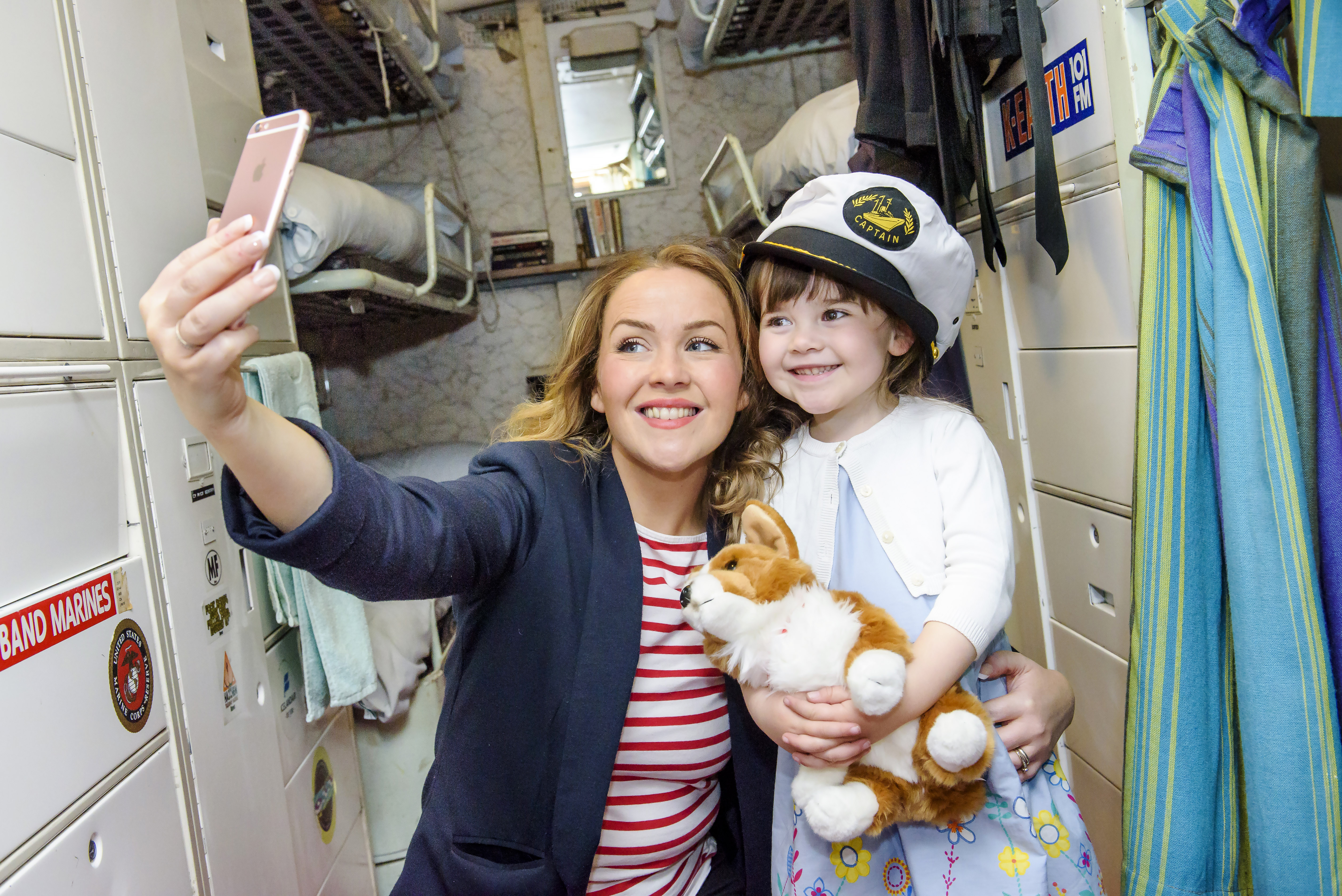 Mother and daughter take a 'selfie' picture together in the ship. The little girl wears a captain's hat and hold a fluffy corgi toy.