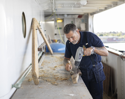 Standing on one of Britannia's decks, a Maintenance man is holding a heat gun and a scraper, removing varnish from a handrail. 