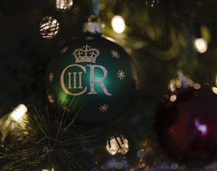A green Christmas bauble with the word CRIII and a crown image printed in gold glitter, hanging on a Christmas tree. 