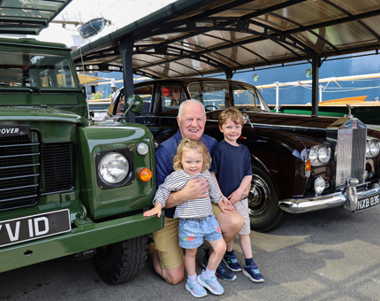 A man with two small children crouch beside the Land Rover and Rolls Royce.
