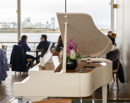 A white grand piano in the Royal Deck Tearoom. There are people sitting at tables in the background