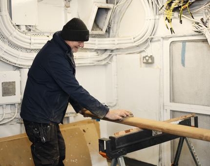 A Maintenance man is sanding a wooden handrail. 