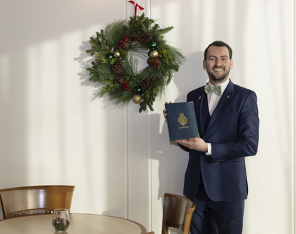 A man standing in the Royal Deck Tearoom aboard Britannia, holding a menu. There is a festive wreath behind him. 
