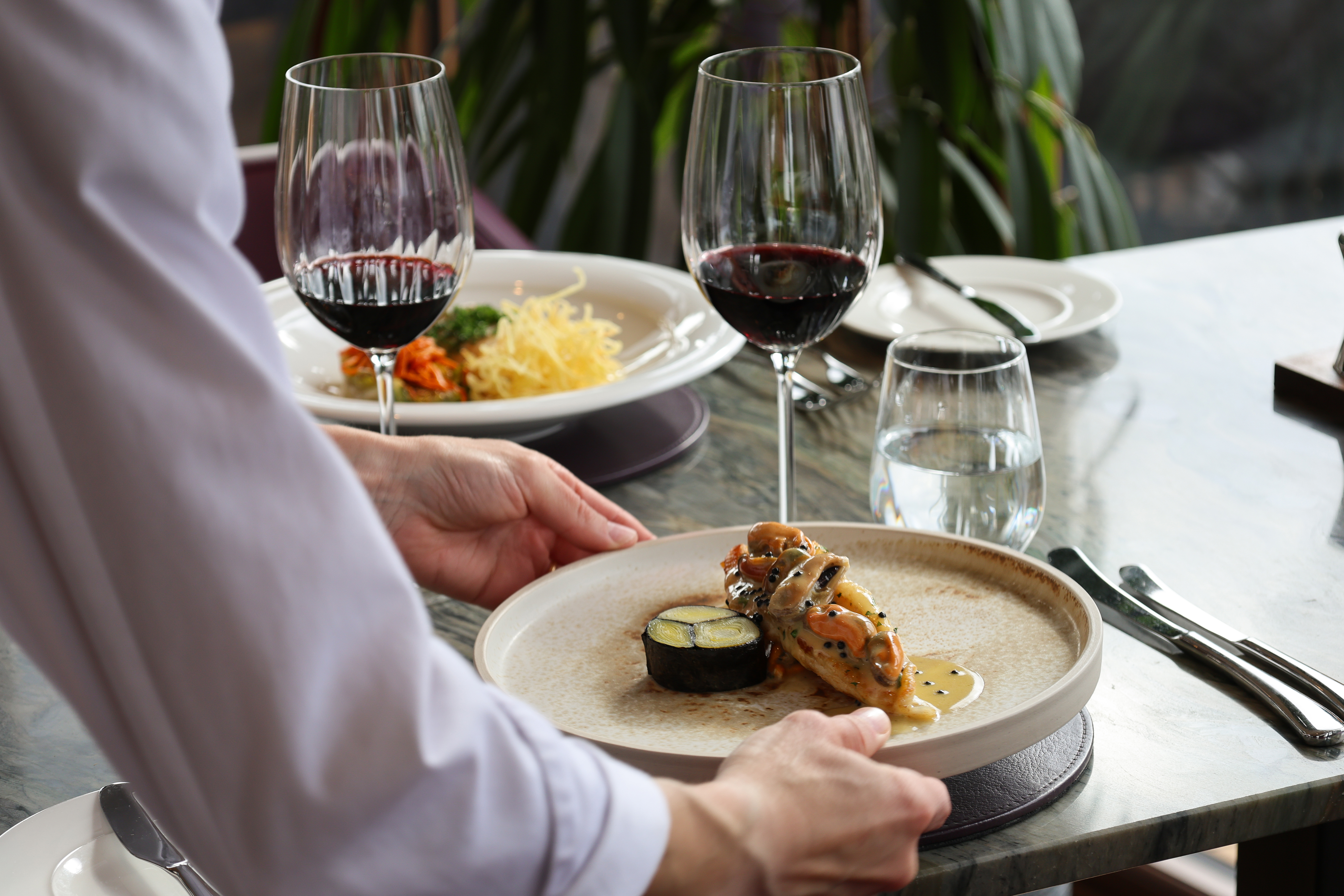 A person is placing a plate onto a table, it has a fish dish on it. There are two glasses of red wine in The Lighthouse Restaurant in Leith. 