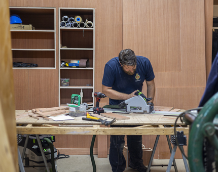 A man is in the workshop cutting wood to build storage. There are various tools on the table next to him.