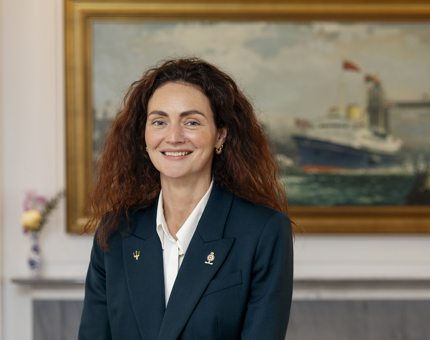 A woman is posing for a photo in front of a painting in Britannia's Drawing Room. 