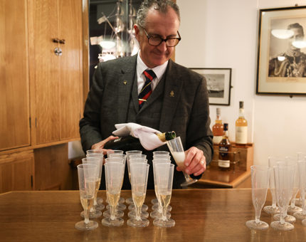 A man wearing a suit is pouring glasses of Champagne in the Wardroom at Britannia. 