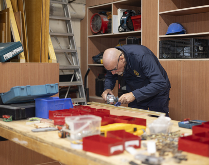 A Maintenance man is bending over a workbench, cutting wood for a door threshold tbar. 