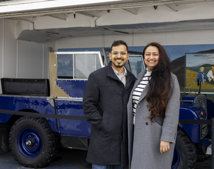 Two visitors posing for a photo by Britannia's onboard Garage, they are standing in front of a blue Land Rover. 