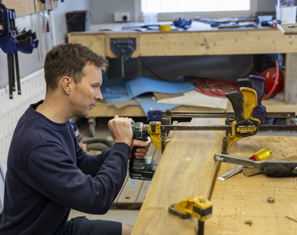 A maintenance man is squatting next to a workbench to repair a handrail. 