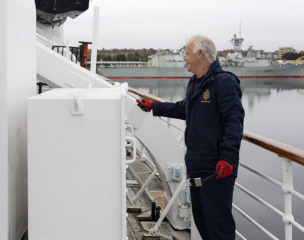 A maintenance man is using a paint roller to apply a coat of paint on deck. A navy ship is in the background. 
