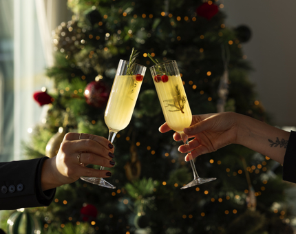 Two hands are clinking glasses of Mimosas in front of a Christmas tree in the Royal Deck Tearoom in Leith. 