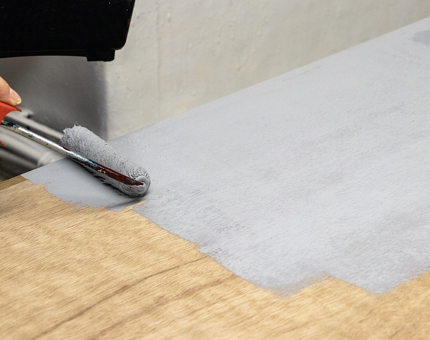 A hand is holding a small paint roller, adding a coat of grey paint to a panel of wood. 