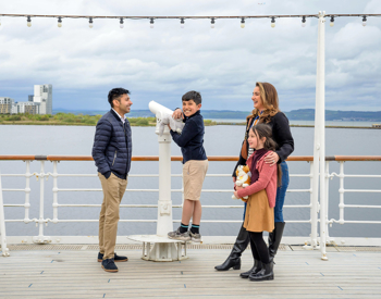 A family of four on the Verandah Deck of Britannia, the small boy is next to a telescope, there is water in the background. 