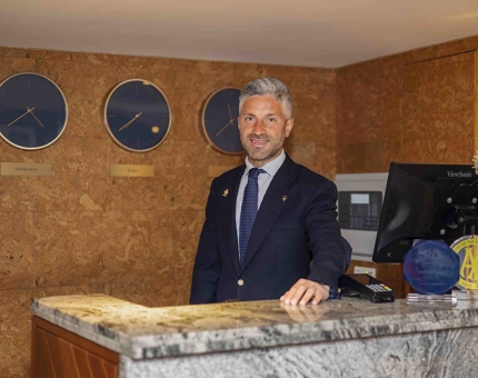 A man posing for a photo at Fingal Hotel's reception. He is standing behind the reception desk, there are three round clocks behind him on the wall. 