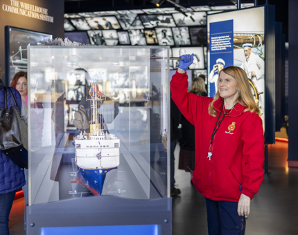 A woman wearing a red Britannia jacket is dusting a glass display case containing a model of The Royal Yacht Britannia. 