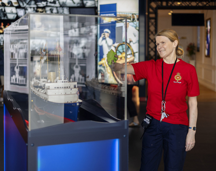 A Housekeeper is holding a cloth, wiping a glass case which holds a replica model of Britannia in the Visitor Centre.