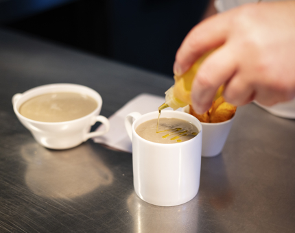 A close up of a chef drizzling oil onto a mug of soup. 