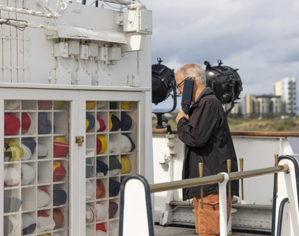 A man is looking at the flag locker on The Royal Yacht Britannia's Bridge while listening to the audio guide handset. 
