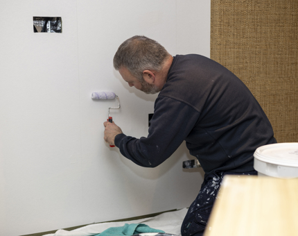 A maintenance team member holding a small roller applying wallpaper paste to a wall. 