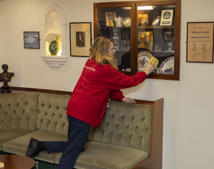 A Housekeeper is kneeling on a green seat and polishing a glass cabinet aboard Britannia in Leith. 