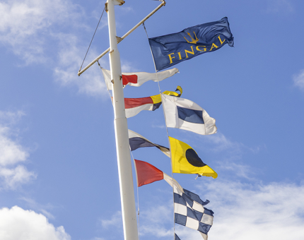 Fingal Hotel's flag pole with colourful flags flying against a blue sky. 