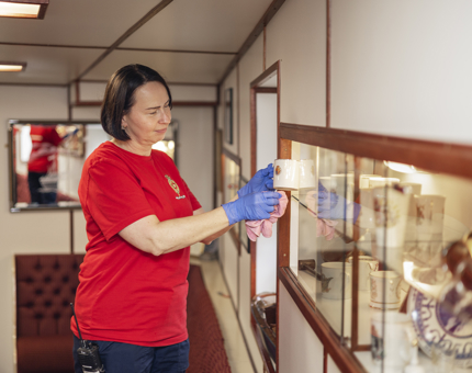 A Housekeeper in Britannia's Unwinding Room is cleaning items in a cabinet. She is holding a mug.