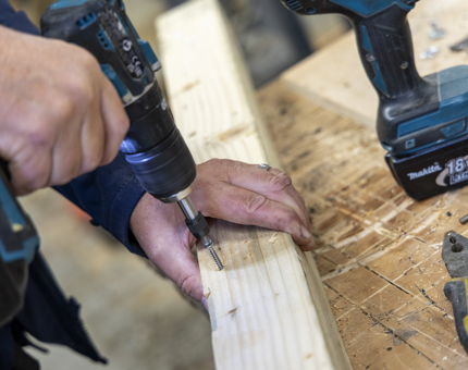 A close up of a hand using a drill, making a hole in a piece of wood. 