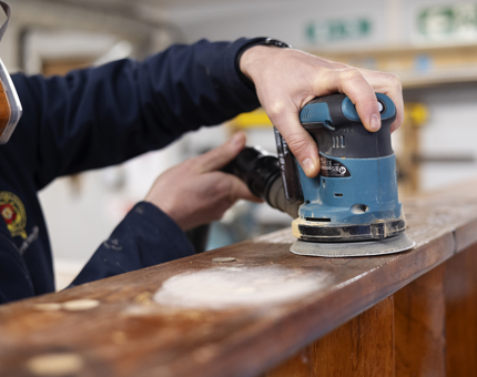 A close up of a hand holding an electric sander, sanding wooden stairs. 