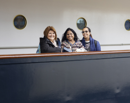 Three women stand behind the railing on one of Britannia's decks in the Port of Leith. 