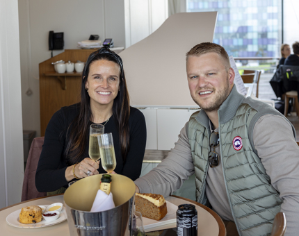 A couple sitting at a table in the Royal Deck Tearoom in Leith holding glasses of Champagne. There is a scone and a slice of  cake on the table next to a Champagne bucket. too. 