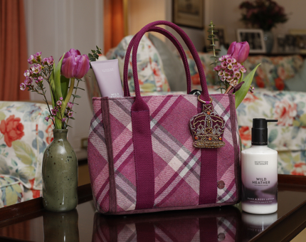 A pink and purple tartan style handbach woth a beaded crown keychain sits on a table with a bottle of Wild Heather hand cream and vases with pink tulips in them. 