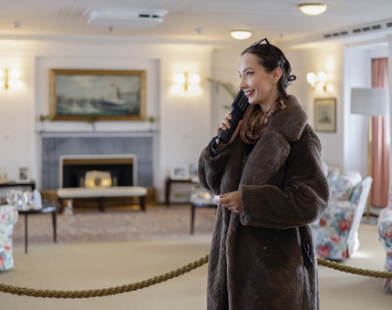 A woman standing in the Drawing Room aboard The Royal Yacht Britannia listening to an audio guide handset.