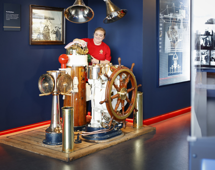 A Housekeeper is in Britannia's Visitor Centre, she is polishing a wood and brass ship's wheel. 