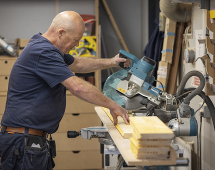 A Facilities Officer is at a workbench, he is cutting wood with a circular saw. 