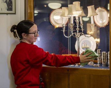A Housekeeper is cleaning a glass cabinet with silver items in it in the Officer's Wardroom aboard Britannia. 