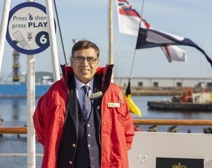 A Visitor Assistant wearing a red jacket posing for a photo. There are flags flying in the background. 