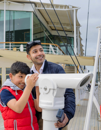 A little boy looking through a telescope at The Royal Yacht Britannia. His Dad is looking on at the view. 