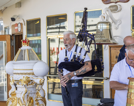 A man playing bagpipes. He is standing behind the decorative replica binnacle.