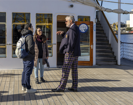 A male Visitor Assistant wearing a tartan uniform is talking to two visitors on the Verandah Deck. 