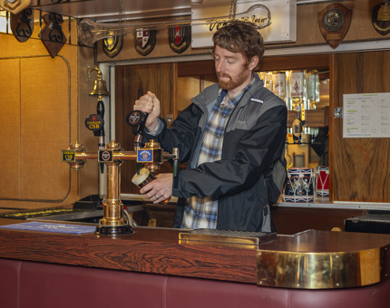 A man stands behind the bar in the Crew's Mess on The Royal Yacht Britannia pretending to pour a pint.