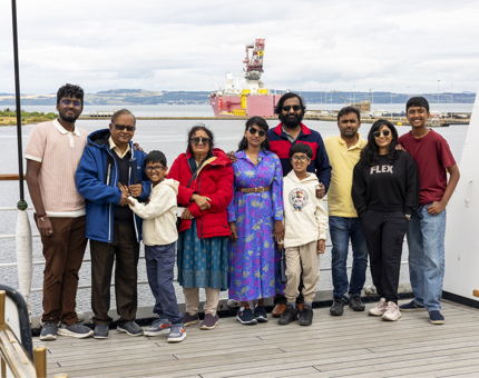 A group of people of various ages are posing for a photo on Britannia's Bridge. A view over the Firth of Forth is in the background. There is a red and white ship. 
