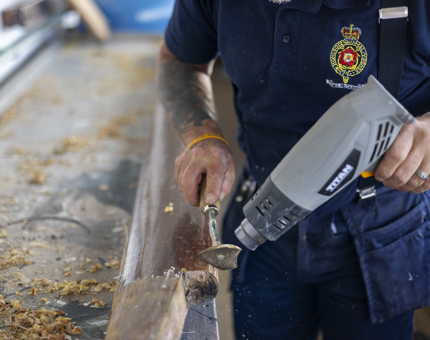 A close up of a hands holding a scraper and a heat gun to remove varnish from a handrail.
