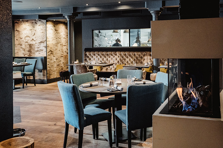 An atmospheric image of a restaurant interior. There is a fireplace in the right hand side, the chairs are covered in blue material. In the background there is a window where you can see chefs cooking. 