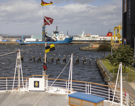 The aft of Britannia. There are colourful dress flags flying in the wind and a view of ships in the Port of Leith in the background.