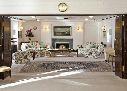 An elegant lounge is viewed through open wooden doors, with two floral sofas facing each other across a wooden coffee table. A fireplace with a ship painting above it sits at the back of the room, framed by wall sconces, while a patterned rug and a vase of flowers complete the refined setting.