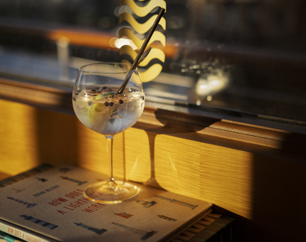 A glass of gin & tonic with a black straw in it is sitting by a window in The Lighthouse Restaurant. Evening sun is shining onto it. 